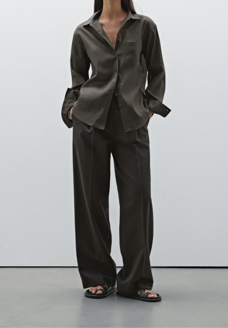 Chemise à boutons vert olive foncé associée à un pantalon ample assorti. Silhouette décontractée, tissu doux et poignets prolongés. Sandales noires.