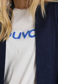 Mörkblå cardigan över en vit t-shirt med blå broderad text. Cardiganen har en mjuk textur och öppen framsida.