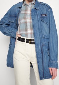 Denim takki keskmises sinises värvitoonis, millel on lukuga sulgemine, rinnakott ja õlavarrepaelad. Kombineeritud ruudulise särgi ja valgete kõrge taljes pükstega.