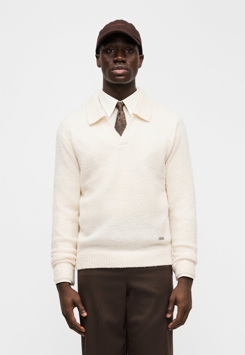Cream sweater with a v-neck and collar, textured fabric, paired with a white shirt, patterned tie, and dark brown trousers. Brown cap.