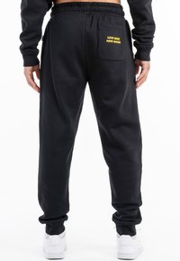 Pantalones de chándal negros con un corte relajado, cinturilla elástica y bolsillo trasero con texto amarillo: "AMOR A LA MÚSICA, ODIO AL RACISMO."