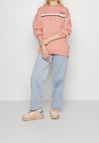 Sudadera rosa con un bolsillo frontal y capucha con cordón. Presenta rayas blancas y marinas en el pecho. Combinada con pantalones de pana azul claro.