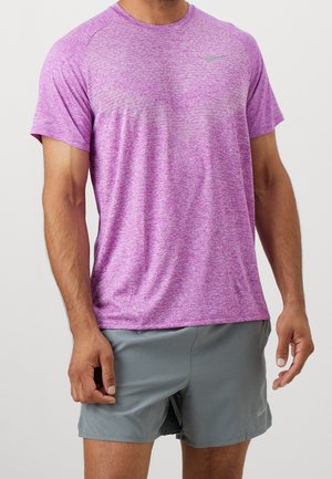 Mann trägt ein kurzärmeliges violettes Sportshirt und graue Shorts, steht mit entspannten Armen an den Seiten.