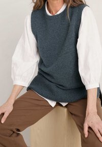 Grauer Strick-Ärmelloser Pullover über einer weißen strukturierten Bluse, kombiniert mit braunen Hosen. Der Pullover hat einen gerippten Saum und eine lockere Passform.