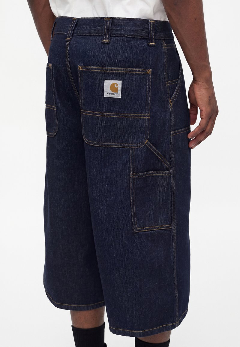 Mørkeblå denimshorts med flere praktiske lommer, synligt Carhartt-logo på baglommen, båret af en person med mørk hud og sorte sokker.