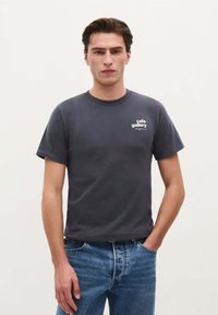 Tricou din bumbac gri închis, cu mâneci scurte, având un imprimeu alb cu logo-ul "cafe gallery" pe zona stângă a pieptului. Purtați cu blugi albaștri.