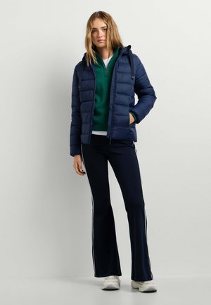 Junge Frau trägt eine marineblaue Steppjacke über einem grünen Zip-up-Pullover, schwarze Hosen mit weißen Streifen und weiße Sneakers, steht vor einer schlichten Wand.