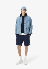Giacca di jeans di colore azzurro chiaro, indossata sopra un hoodie blu navy e una maglietta bianca. Pantaloni corti blu navy, sneakers bianche e un berretto blu coordinato con logo.