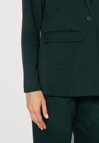 Main de femme reposant à côté d'un blazer cintré vert foncé avec poche à rabat et pantalon assorti sur fond clair.