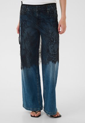 Weit geschnittene blaue Jeans mit schwarzer floraler Spitzenüberlagerung von der Taille bis zur Mitte des Oberschenkels, getragen mit schwarzen Zehensandalen auf hellem Hintergrund.