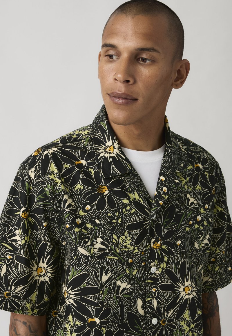 Uomo con capelli corti che indossa una camicia nera decorata con grandi fiori bianchi e gialli sopra una maglietta bianca, che guarda leggermente verso destra.