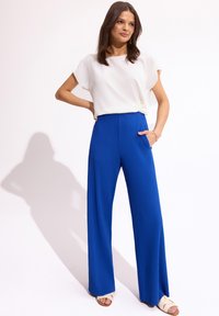 Biała bluzka o luźnym fasonie z detalem w postaci węzła, zestawiona z długimi, plisowanymi spodniami w jasnym niebieskim kolorze oraz beżowymi klapkami.