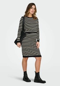 Robe en maille à rayures verticales noires et crème, encolure ronde, manches longues, forme cintrée, et ourlet côtelé. Associée à des bottines noires.