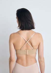 Beige BH med kryssende stropper og krok-og-løkke-lukking. Glatt tekstur, justerbare stropper, minimalistisk design.