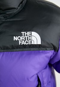 Lilla pufferjakke med svart overdel, med "The North Face" logo i hvit brodering. Glatt tekstur med en lett glans.