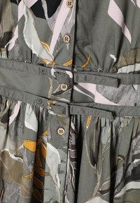 Detailní záběr na šaty z látky s květinovým vzorem, s knoflíkovými detaily a dvěma ladícími látkovými pásky zapnutými v pase.