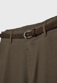 Pantalon marron avec une ceinture en daim marron foncé et une boucle argentée, présenté de la taille au haut de la cuisse sur un fond clair et uni.
