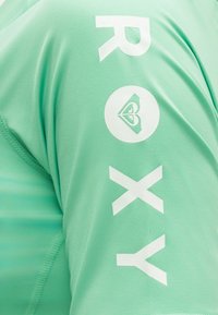 Πράσινο ύφασμα με λευκή επιγραφή "ROXY" και κυκλικό λογότυπο με στυλιζαρισμένη καρδιά σε μανίκι ενδύματος.