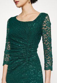 Femme portant une robe en dentelle vert foncé avec des manches trois-quarts et un détail froncé à la taille, associée à des boucles d'oreilles dorées.