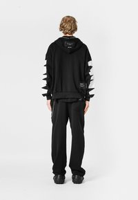 Sudadera negra oversized con elementos gráficos blancos en las mangas, combinada con pantalones negros y calzado negro de plataforma, contra un fondo simple.