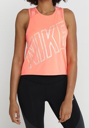 Kvinde iført en ærmeløs koralfarvet Nike-top kombineret med sorte træningsleggings, stående foran en ensfarvet baggrund.