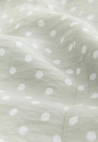 Hellgrüner Stoff mit subtiler Textur, versehen mit gleichmäßig verteilten weißen Punkten und sichtbaren Nahtdetails.