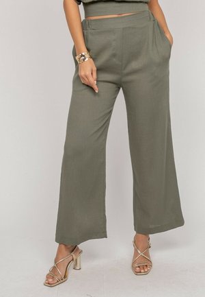 Femme portant un pantalon large kaki, un bracelet manchette doré et des sandales à talons hauts dorées à lanières, debout les mains dans les poches.