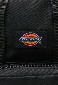 Mustrikangast must kangas, millel on kudumine, millel on sinine ja punane "Dickies" logo mustal lipikul, õmmeldud sirge äärega ja lukuga.