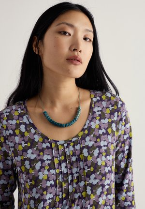 Ljubičasti top s cvjetnim uzorkom u nijansama plave i zelene, s izrezom u obliku slova U i detaljima na nabore. Teksturirana plava ogrlica upotpunjuje izgled.