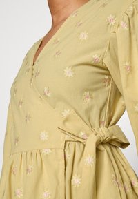 Vestido amarelo claro com um design envolvente, apresentando delicados bordados florais em rosa e branco. Inclui um detalhe de laço na cintura e mangas bufantes.