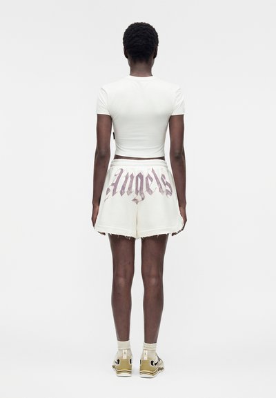 Άτομο με την πλάτη, φοράει λευκό κοντό μπλουζάκι, λευκά σορτς με το κείμενο "Angels" σε γοτθική γραμματοσειρά, μπεζ κάλτσες και μπεζ αθλητικά παπούτσια.