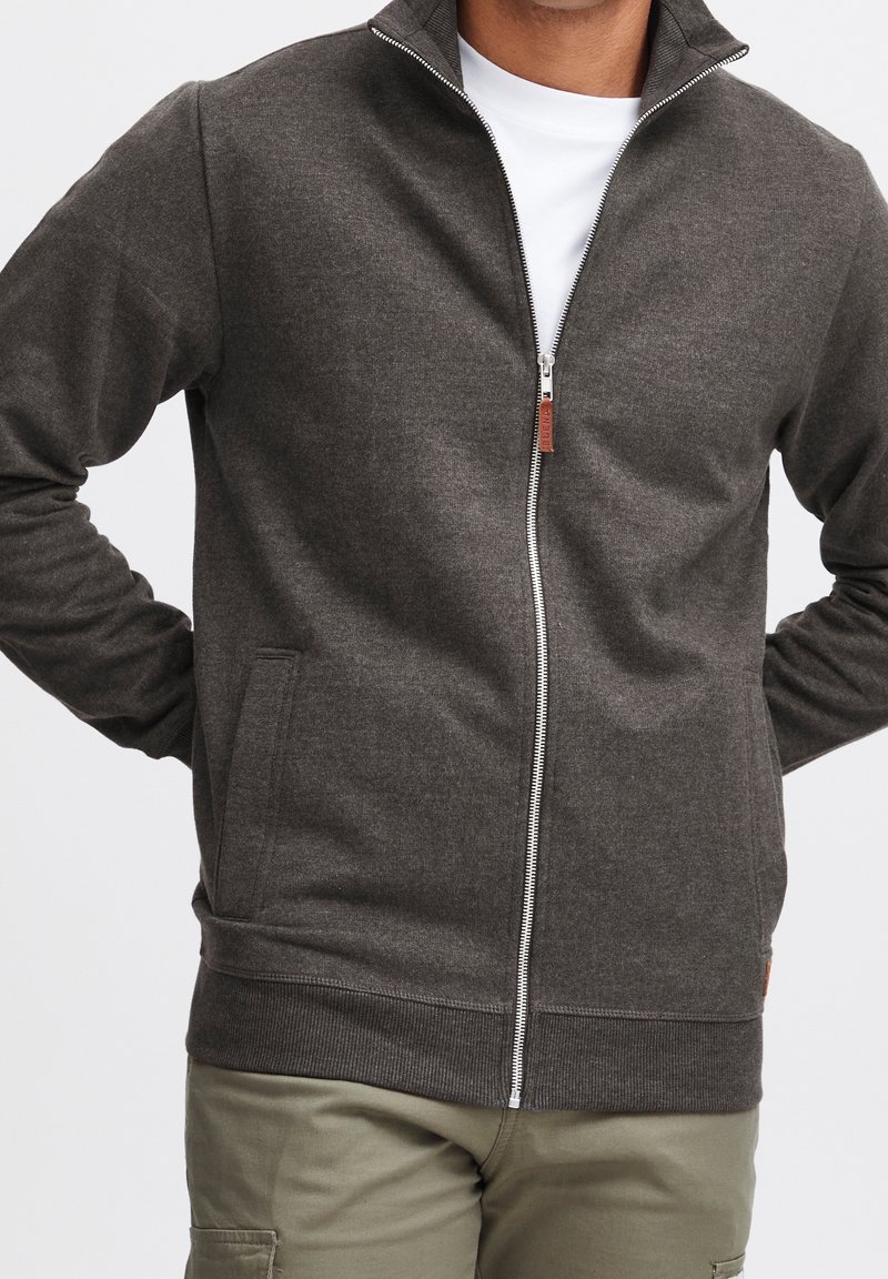 Veste gris zippée en tissu doux, avec un col montant, deux poches latérales et un détail en cuir près de la fermeture éclair.