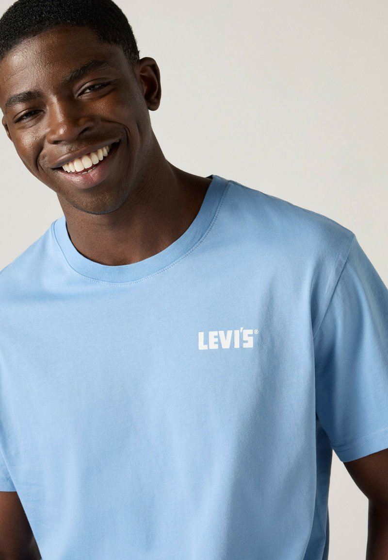 Lächelnder Mann, der ein hellblaues Levi's T-Shirt vor einem neutralen, einfarbigen Hintergrund trägt und leicht nach unten schaut.