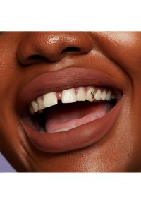 Matts brun läppstift på fylliga läppar, som visar vita tänder med en liten lucka. Huden har en jämn, djup ton, och en guld tandaccent syns.