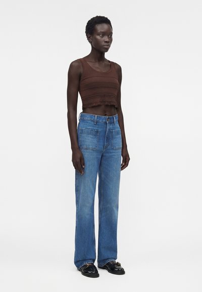 Ψηλό άτομο στέκεται, φορώντας ένα καφέ cropped μπλουζάκι χωρίς μανίκια, τζιν με ψηλή μέση και μπροστινές τσέπες, και μαύρα loafers, πάνω σε λευκό φόντο.