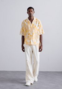 Chemise à manches courtes jaune avec un motif abstrait, associée à un pantalon blanc ample comportant un détail imprimé subtil. Les deux vêtements ont une coupe décontractée.