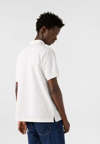 Polo bianco con maniche corte, fessure laterali, realizzato in cotone, caratterizzato da un colletto classico e un design semplice, indossato con jeans blu scuro.