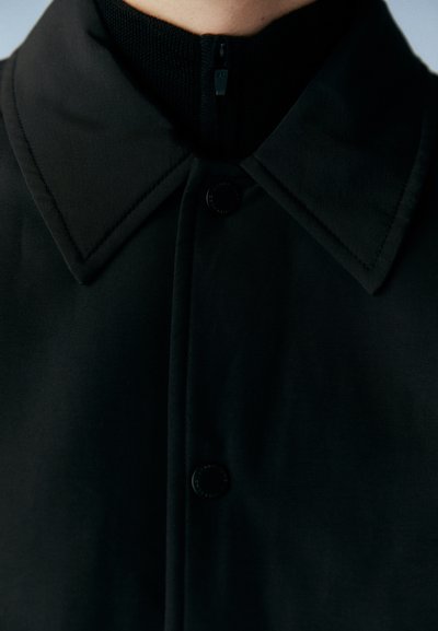 Μαύρο μπουφάν με κολάρο που διαθέτει υφή, φερμουάρ κλειστού κολάρου και κουμπί μπροστά, με λείος, ματ φινίρισμα.