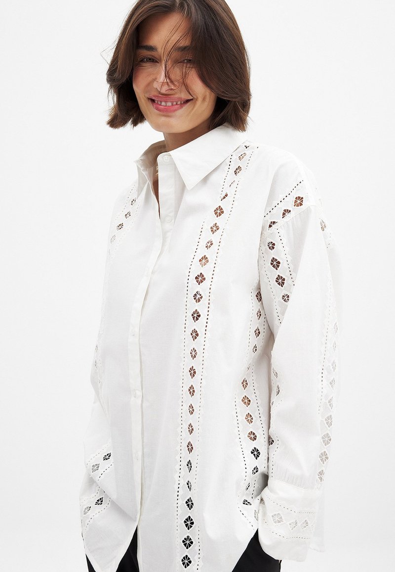 Camicia bianca con bottoni, caratterizzata da un'intricata ricamo bianco lungo i lati, con una vestibilità morbida e maniche lunghe. Design del colletto semplice.