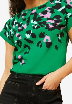 Chemise verte à manches courtes avec des taches de léopard noires et roses portée avec un pantalon noir taille haute, main dans la poche droite.