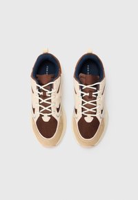 Et par brune og beige net sneakers med hvite snørebånd, sett ovenfra på en ensfarget lys grå bakgrunn.