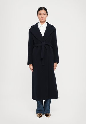 Donna in piedi con un lungo cappotto nero con cintura sopra una camicia bianca e jeans blu, abbinati a scarpe a punta con stampa leopardata, su uno sfondo bianco.