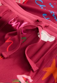 Close-up of red fabric pants with a tied waistband drawstring and colorful abstract text and star patterns.