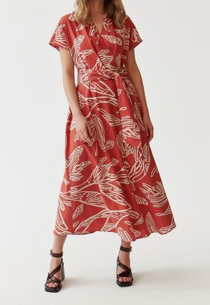 Femme portant une robe portefeuille couleur rouille avec des motifs abstraits crème en forme de feuilles et des sandales noires à talons hauts sur un fond uni.
