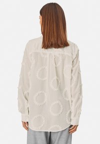 Long-sleeve, cream shirt featuring textured, circular embroidery, soft fabric, button cuffs, and a relaxed fit. Back view displayed.