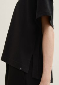 Schwarzes T-Shirt aus weichem Stoff, mit einem lockeren Schnitt, seitlichen Schlitzen und einem kleinen Logotag am Saum. Kurze Ärmel und schlichtes Design.