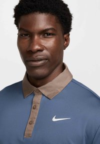 Nike polo krekls ar zilu korpusu un bēšām apkaklēm. Tam ir pogām aprīkota apmale un balts swoosh logs uz krūšu daļas. Gluda tekstūra.