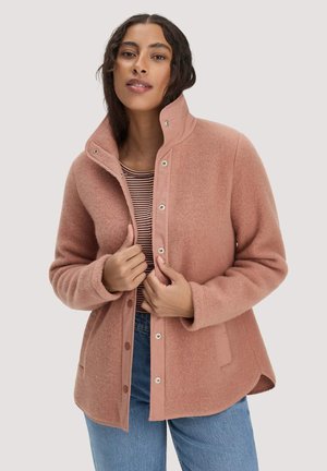 Woman wearing a dusty pink buttoned jacket over a striped shirt and blue jeans, adjusting the jacket collar and front.