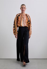 Geprinte blouse met lange mouwen in oranje en zwarte patronen, gecombineerd met hoge zwarte wijde broek met splitten aan de zijkant en gerimpelde details.
