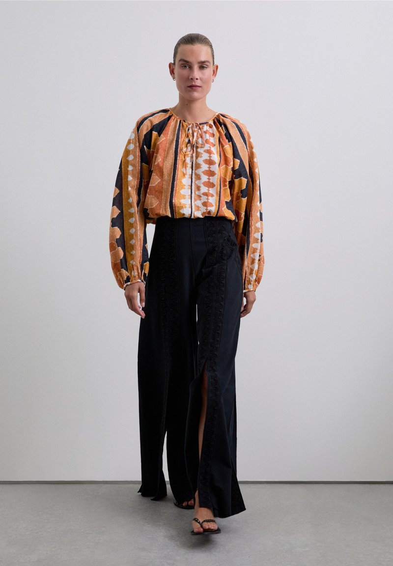 Geprinte blouse met lange mouwen in oranje en zwarte patronen, gecombineerd met hoge zwarte wijde broek met splitten aan de zijkant en gerimpelde details.